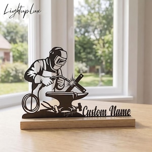 May include: A black metal silhouette of a welder wearing a helmet and gloves, working on a metal piece over an anvil. The design is mounted on a rectangular wooden base and features the text Custom Name in a script font on the right side.