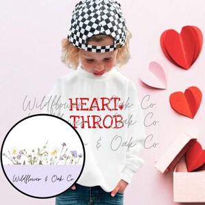 Op de afbeelding: Een kind draagt een witte trui met de rode tekst "HEART THROB". Het kind draagt een zwart-wit geruite pet. Rode en roze papieren harten zijn gerangschikt op een roze achtergrond, sommige harten in een kleine doos.