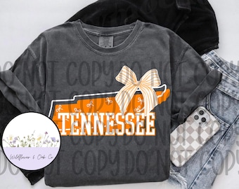 Może przedstawiać: Ciemnoszara koszulka z długim rękawem z pomarańczowo-białym stanem Tennessee i kokardą. Słowo "TENNESSEE" jest wydrukowane na obrysie stanu. Obok koszulki znajduje się telefon z kratkowanym etui i dżinsy.