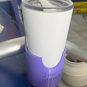 May include: A white tumbler with a clear lid sits inside a purple cup holder attachment. The purple holder features a cutout design and the embossed text TRIPLE DIVIDE with a wavy line graphic. The assembly is mounted on a silver metal bar.