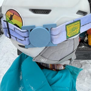 May include: White ski helmet with a blue and white goggle strap, a rainbow sticker, and a yellow sticker that says "SMITH Craylor" with a sun and mountain graphic. The helmet has a blue camera mount.