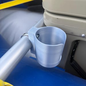 May include: A light blue, 3D-printed cup holder attached to a silver metal bar. The cup holder is mounted on a blue and gray surface, with a yellow strap visible. The cup holder is designed to hold a beverage.