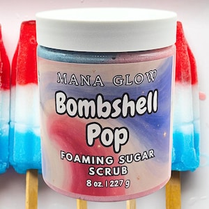 May include: A cylindrical jar of Mana Glow Bombshell Pop foaming sugar scrub with a white lid. The label features a red, white, and blue swirl design with the text "Mana Glow Bombshell Pop Foaming Sugar Scrub 8 oz. | 227 g" printed on the front.