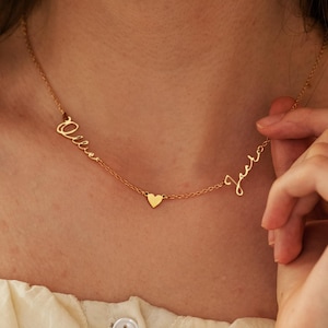 Op de afbeelding: Een goudkleurige ketting met de namen "Olivia" en "Jack" in cursief schrift. Een kleine hartvormige bedel staat tussen de namen. De ketting wordt om de nek van een persoon gedragen.