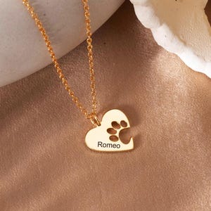 May include: Gold-coloured necklace with a heart-shaped pendant. The pendant has a paw print and the name "Romeo" engraved. The necklace is displayed on a neutral background.