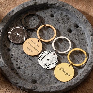 May include: A selection of personalised keyrings featuring map designs and round tags. Some tags have the text "I love you", while others display names, coordinates, and a date. The keyrings are in gold, silver, and black.