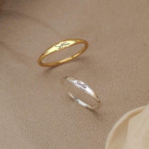 Puede incluir: Dos anillos finos de metal, uno dorado y otro plateado, cada uno con una banda lisa y redondeada con una superficie superior plana. Ambos anillos están grabados con el nombre Stella en una delicada letra cursiva negra en el centro.