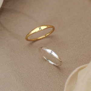 Könnte beinhalten: Zwei dünne Metallringe mit glatter, polierter Oberfläche. Ein Ring ist goldfarben und trägt den Buchstaben R auf seiner flachen Oberfläche eingraviert. Der zweite Ring ist silberfarben und trägt den Buchstaben S eingraviert.