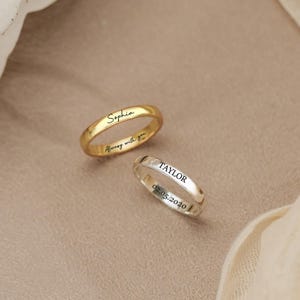 May include: Two thin metal rings, one gold-toned and one silver-toned. The gold ring is engraved with the name Sophia and the phrase Always with you. The silver ring is engraved with the name TAYLOR and the date 02.05.2020 in a sans-serif font.