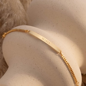 May include: A gold bracelet with the name "MARIA" engraved on a rectangular plate. The bracelet has a curb chain design. The bracelet is displayed on a neutral background.