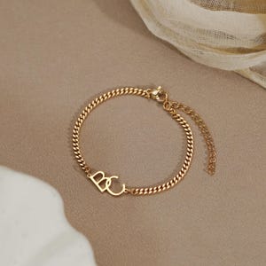 Könnte beinhalten: Ein goldfarbenes Kettenarmband mit zwei ineinander verschlungenen Großbuchstaben, B und C, als zentrales Element. Das Armband besteht aus einer feinen Panzerkette und verfügt über eine verstellbare Verlängerungskette mit Karabinerverschluss.