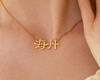 Peut inclure: Un collier doré avec des caractères chinois est porté autour du cou. Le collier est sur une chaîne délicate. La personne porte un haut crème et une montre avec un bracelet argenté.