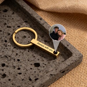 Op de afbeelding: Een goudkleurige rechthoekige sleutelhanger met de gegraveerde tekst "Always with you". De sleutelhanger heeft een cirkelvormige ring en een klein zwart accent. Een kleine cirkelvormige afbeelding van een stel staat op de achtergrond.