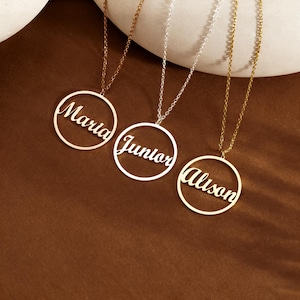 May include: Three personalized name necklaces. Each necklace features a name cut out within a circular pendant. The names are "Maria", "Junior", and "Alison". The necklaces are gold and silver in color, with delicate chains.