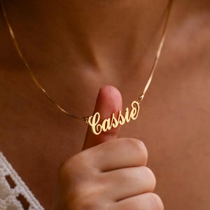 May include: A gold-coloured necklace featuring the name "Cassie" in cursive lettering. The necklace hangs from a fine chain. The name is written in an elegant font. The necklace is held up by a finger.
