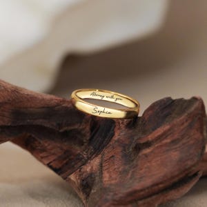 Op de afbeelding: Een gepolijste gouden ring met de gegraveerde naam Sophia en de tekst Always with you in een zwart cursief lettertype. De ring heeft een glad, afgerond buitenoppervlak en een reflecterende metalen afwerking met duidelijk leesbare letters.