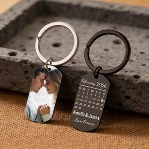 Könnte beinhalten: Zwei personalisierte Schlüsselanhänger. Einer zeigt ein Foto eines Paares, der andere einen Kalender für Februar 2026 mit den Namen "Amelia & James" und den Worten "Love Forever". Beide Schlüsselanhänger haben Metallringe.
