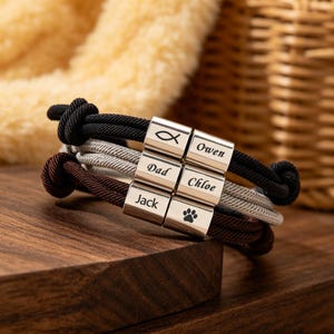 May include: A personalised bracelet with black, grey, and brown cords. The bracelet has silver-coloured beads engraved with names and symbols. The names "Owen", "Dad", "Chloe", and "Jack" are visible, along with a fish symbol and a paw print.