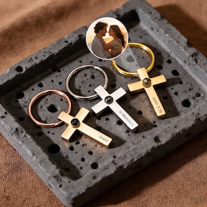 May include: Three cross-shaped keyrings in gold, silver, and rose gold, each with a black gem. The keyrings are engraved with the words "Grace", "In Memory L", and "John 3:16". A small photo of a couple is in the background.