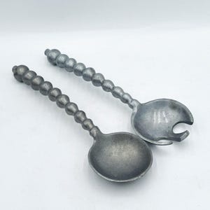 May include: Two vintage silver-toned salad servers. The handles are adorned with a series of small, round beads. One server is a spoon, and the other has a fork-like design. The servers are set against a white background.