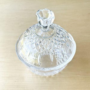 May include: A clear, decorative glass candy dish with a lid. The dish has a diamond pattern and a flower-shaped handle on the lid. The dish is sitting on a light-colored wooden surface.