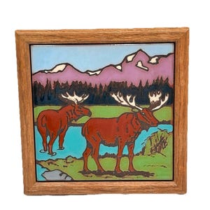 May include: A framed ceramic tile with a colorful illustration of two moose standing in a lake with mountains in the background.