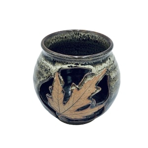 May include: A handcrafted ceramic pot with a dark brown and tan glaze. The pot features a cut-out leaf design on the side. The interior is dark, and the rim has a textured appearance. A decorative piece for the home.