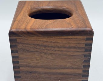 May include: A square tissue box cover crafted from dark brown wood with a visible grain pattern. The structure features interlocking finger joints at the corners, creating a distinct grid-like pattern of alternating light and dark wood segments.