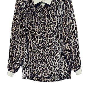 May include: A long-sleeved button-down shirt with a leopard print pattern. The shirt features a cream-colored collar and cuffs. The leopard print consists of brown and black spots on a tan background. The shirt has a classic, vintage style.