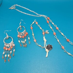 May include: A jewelry set featuring a silver chain necklace, earrings, and bracelet. The earrings and necklace have coral-colored beads and white pearl-like beads. The bracelet has a silver rose charm and a white leather cord.