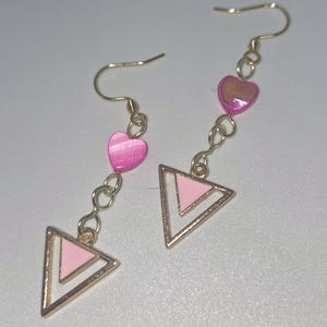 May include: A pair of dangle earrings with gold-colored hooks and chains. Each earring features a pink heart-shaped bead and a pink and white triangle pendant. The earrings are designed with a delicate and feminine aesthetic.