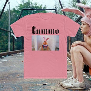 Op de afbeelding: Een roze t-shirt met het woord "Gummo" in gotisch lettertype. Het shirt heeft een afbeelding van een persoon met konijnenoren die op een toilet zit. De persoon draagt ook konijnenoren.
