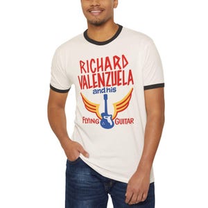 May include: Cream-colored ringer t-shirt with black trim featuring the text "RICHARD VALENZUELA and his FLYING GUITAR" in red and blue. The design includes a blue guitar with yellow wings. The shirt is worn by a person.