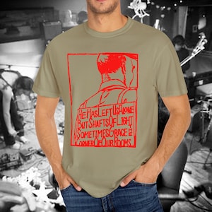 May include: A tan t-shirt with a red graphic design. The design features a silhouette of a person and the text "HE HAS LEFT US ALONE BUT SHAFTS OF LIGHT SOMETIMES GRACE CORNER OF OUR ROOMS."