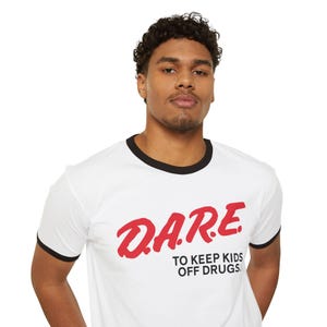 Puede incluir: Camiseta blanca con ribetes negros en el cuello y las mangas. La camiseta presenta la palabra "D.A.R.E." en rojo, con el texto "TO KEEP KIDS OFF DRUGS" debajo en negro.