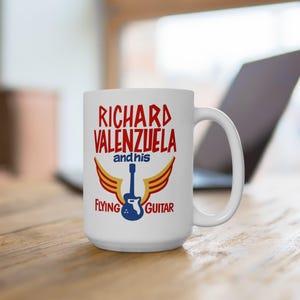 May include: White ceramic mug featuring the text "RICHARD VALENZUELA and his FLYING GUITAR" in red and blue. The design includes a blue guitar with yellow and red wings. The mug has a large handle.