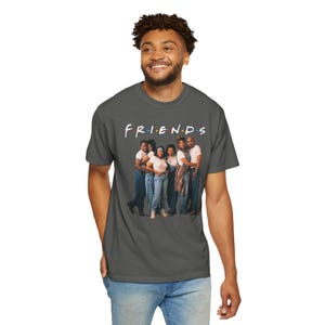 Könnte beinhalten: Ein anthrazitfarbenes T-Shirt mit dem Wort "FRIENDS" in Weiß, das "I" in Rot. Das Shirt zeigt ein Gruppenfoto von sechs Personen. Die Personen tragen weiße und blaue Kleidung. Das Shirt besteht aus einem weichen Material.