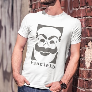 May include: White t-shirt featuring a gray graphic of a stylized face within a rectangular frame, with the text "fsociety" below. The design is printed on a short-sleeved shirt.