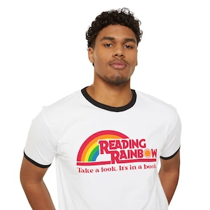 Pode incluir: Camiseta branca com acabamento preto com o logotipo "Reading Rainbow". O logotipo inclui um arco-íris, as palavras "Reading Rainbow" em vermelho e o slogan "Take a look. It's in a book."