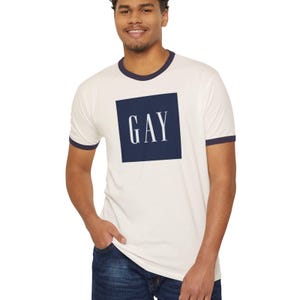 以下が含まれることがあります： クリーム色のリンガーTシャツ。ネイビーブルーのトリムと、ネイビーブルーの正方形の中に白いブロック体で「GAY」の文字がプリントされています。半袖で、柔らかい素材で作られています。