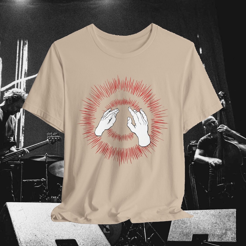 以下が含まれることがあります： ベージュのTシャツ。デザインは、ジェスチャーをする2つの白い手と、赤いバーストパターンで構成されています。Tシャツのニュートラルな色合いは、汎用性があります。グラフィックは前面の中央に配置されています。