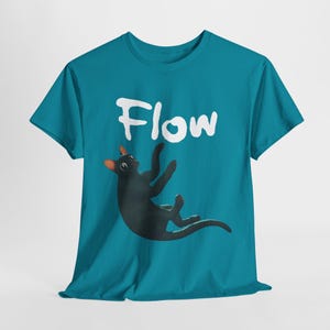 Op de afbeelding: Teal t-shirt met een zwarte kat in een speelse pose en het woord "Flow" in witte letters. De kat heeft oranje oren en lijkt te spelen. Het t-shirt is gemaakt van zacht materiaal.
