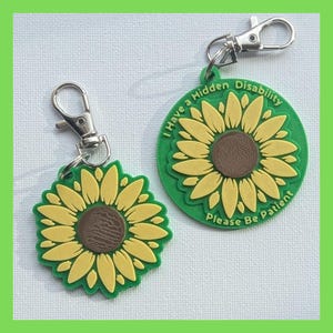 Puede incluir: Dos llaveros de girasol verde con anillas de plata. El llavero m&aacute;s grande tiene el texto "I Have a Hidden Disability Please Be Patient".