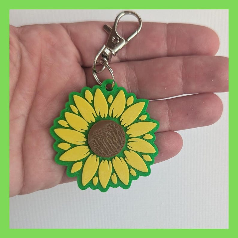 Puede incluir: Un llavero de girasol verde y amarillo con un centro marr&oacute;n. El llavero tiene un cierre de metal plateado.