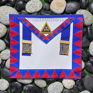 Puede incluir: Delantal masónico blanco con ribetes azules y rojos. El delantal presenta un peto azul con un emblema dorado y flecos de cadena dorada. El delantal está bordeado con triángulos rojos y el texto "handmadeMasonShop".