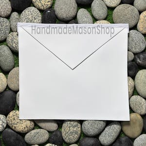 May include: A white Masonic apron with a pointed bottom edge and a white band at the top. The text "HandmadeMasonShop" is printed across the top. The apron is displayed on a bed of rocks and green grass.