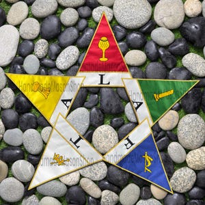 Può includere: Una disposizione a forma di stella di bandiere triangolari colorate con bordi dorati, su uno sfondo di ciottoli ed erba. Ogni bandiera presenta un colore e un simbolo diversi, tra cui un calice, un fascio di grano e una tromba. Le bandiere hanno il testo "HandmadeMasonShop".