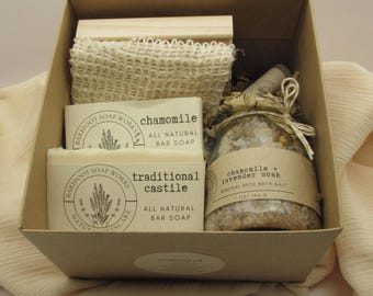 Puede incluir: Una caja de regalo de cartón que contiene dos pastillas de jabón rectangulares de Barefoot Soap Works etiquetadas como chamomile y traditional castile, un frasco de sales de baño chamomile and lavender soak de 7 oz/196 g y una esponja beige.