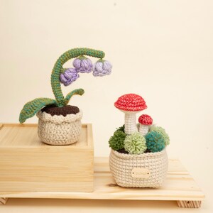 Puede incluir: Dos decoraciones de plantas de ganchillo. Una presenta un tallo verde con flores moradas en una maceta beige. La otra tiene figuras de hongos rojos y blancos en una maceta beige con detalles de pompones verdes y azules. Ambas están sobre una superficie de madera.