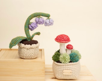 May include: Two crocheted plant decorations. One features a green stem with purple flowers in a beige pot. The other has red and white mushroom figures in a beige pot with green and blue pom-pom accents. Both are on a wooden surface.
